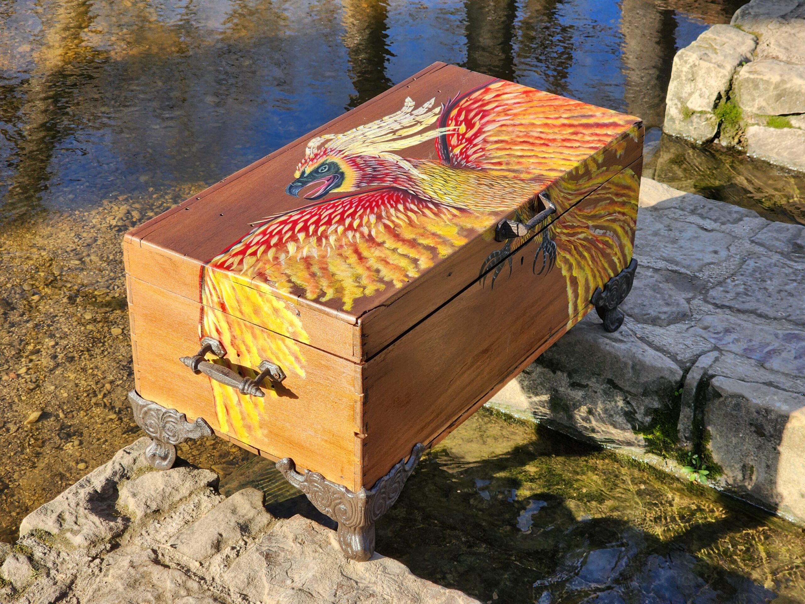 This is an antique walnut wood chest/box that I repurposed into a trinket box. I had to sand it off, and then I handpainted the design of the phoenix over the box with acrylic paint. Then, the box was varnished to protect it from external debris. The feet and the handles are made of cast iron and were professionally installed on the box. I decided to leave the inside of the box untouched to show the age of the box and to give it character.